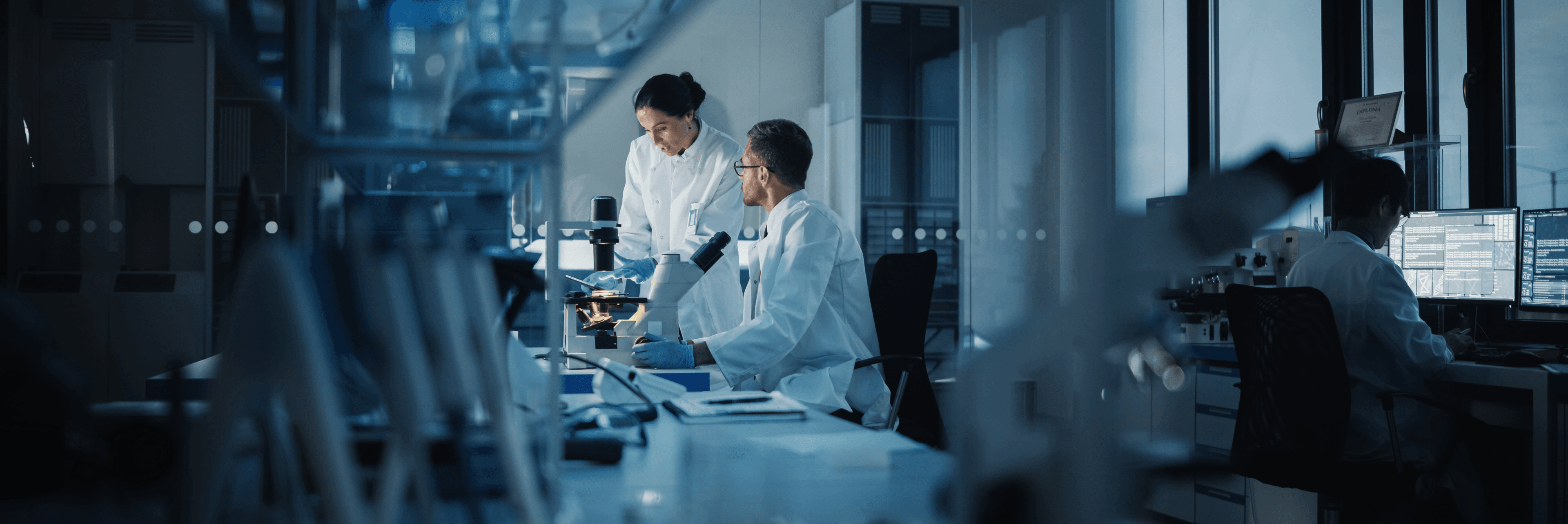Two medical researchers in white lab coats collaborating over a microscope in a modern blue-lit laboratory, with a third researcher working at monitors in the background
