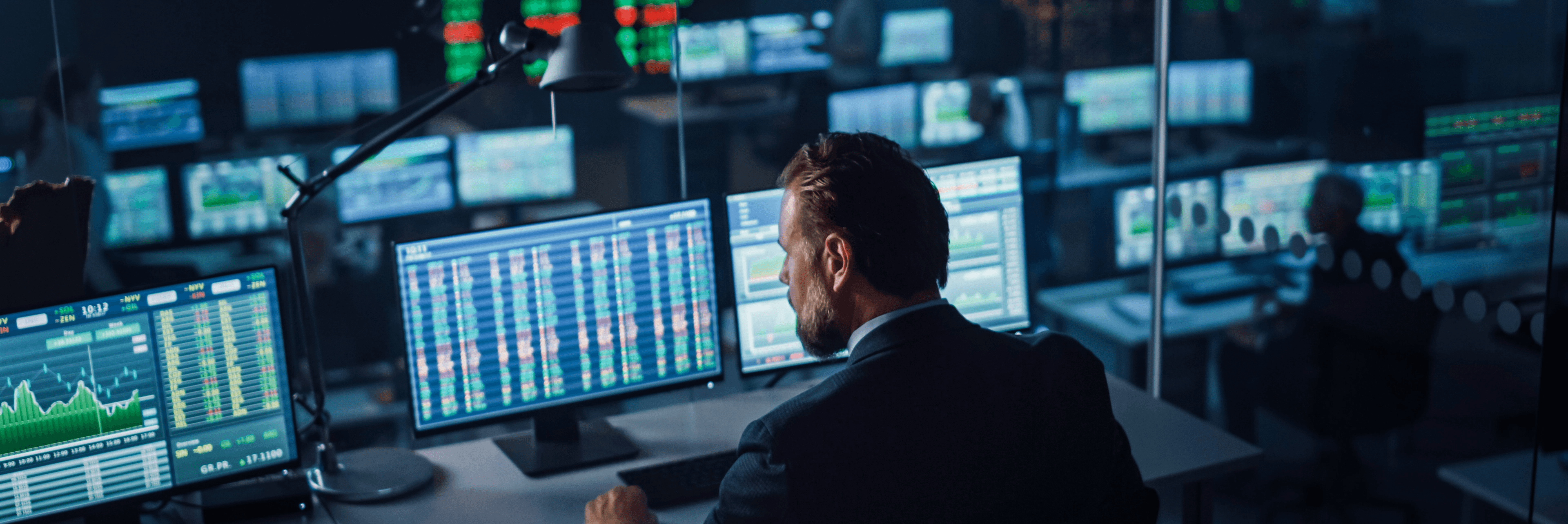Financial trader monitoring multiple screens displaying live stock market data, trading charts, and price feeds in a dark professional trading floor environment