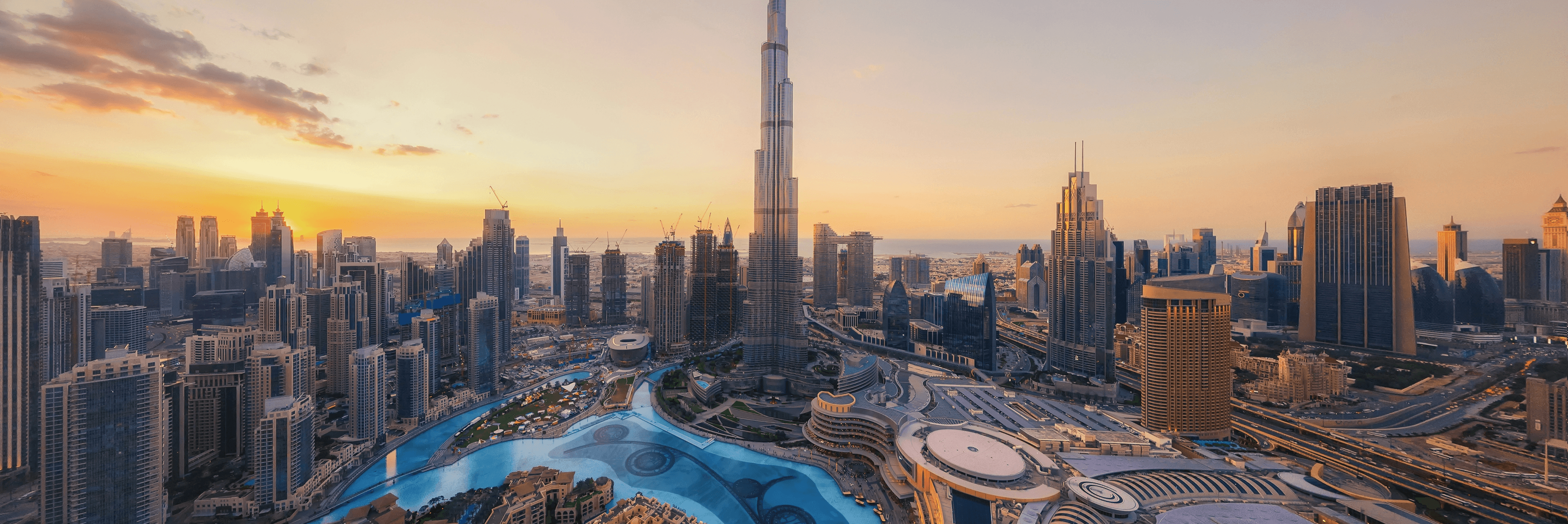 Aerial panoramic view of the Dubai skyline at sunset with the Burj Khalifa towering over the city, the Dubai Fountain lake, and the Persian Gulf visible in the golden sky background